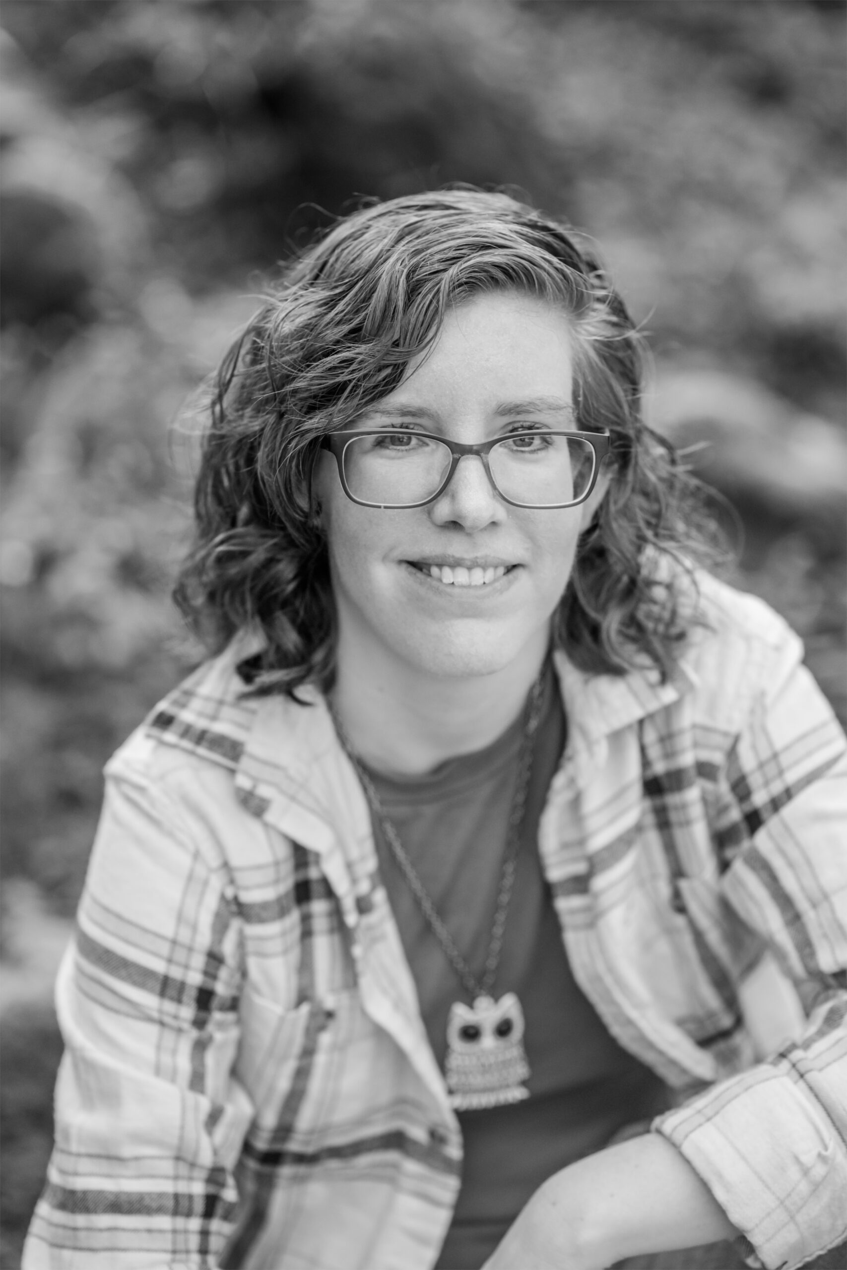A black and white photo of Jasmine from the waist up. Jasmine is a white genderfluid person with curly shoulder-length hair wearing glasses, a plaid button-up shirt, and an owl pendant.